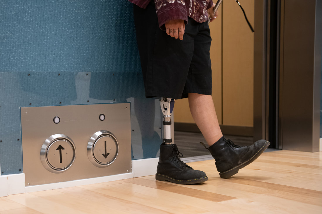 Lower body crop of an Indigenous Two-Spirit person with a prothestic leg standing next to an open elevator and its floor-located call buttons. The word “free” can be seen tattooed across their knuckles, and the person is also holding a cane.